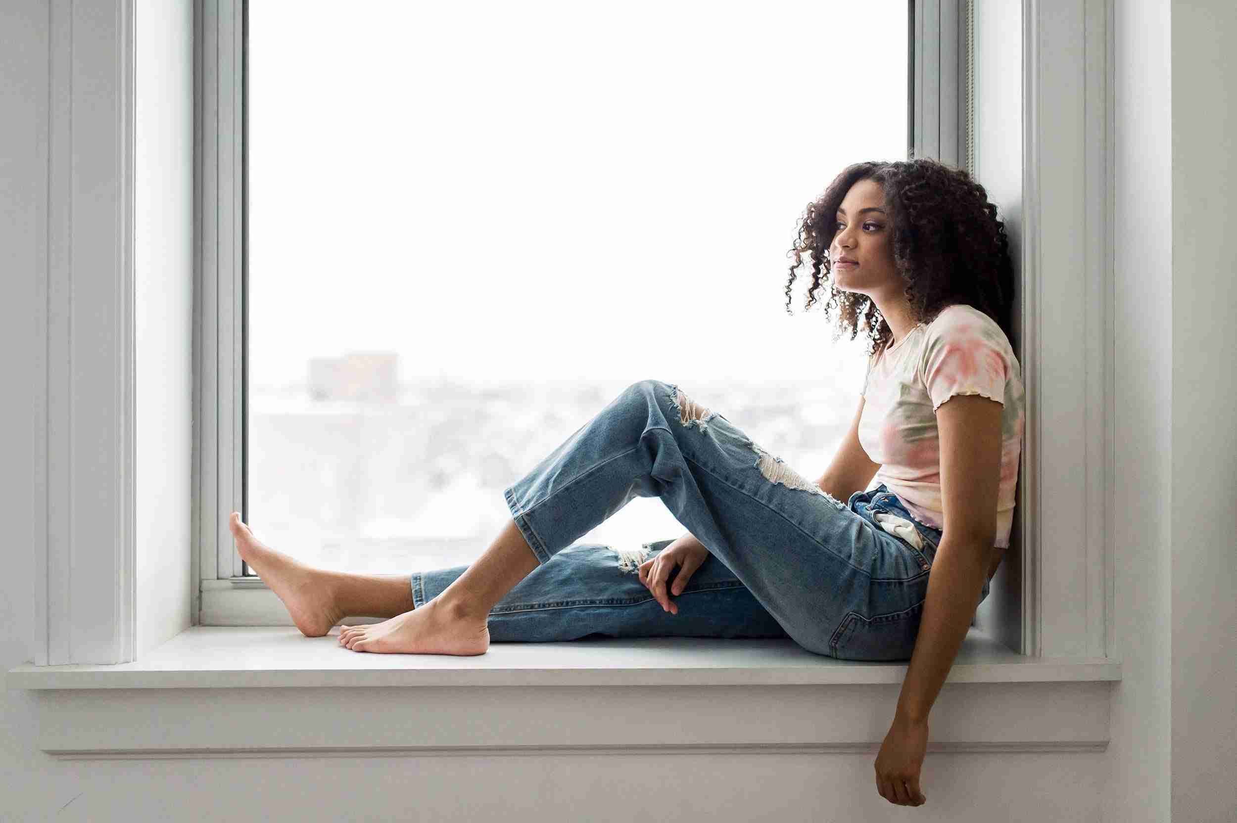 Girl wearing jeans and a tie dye t-shirt sitting on the inside ledge of a window.