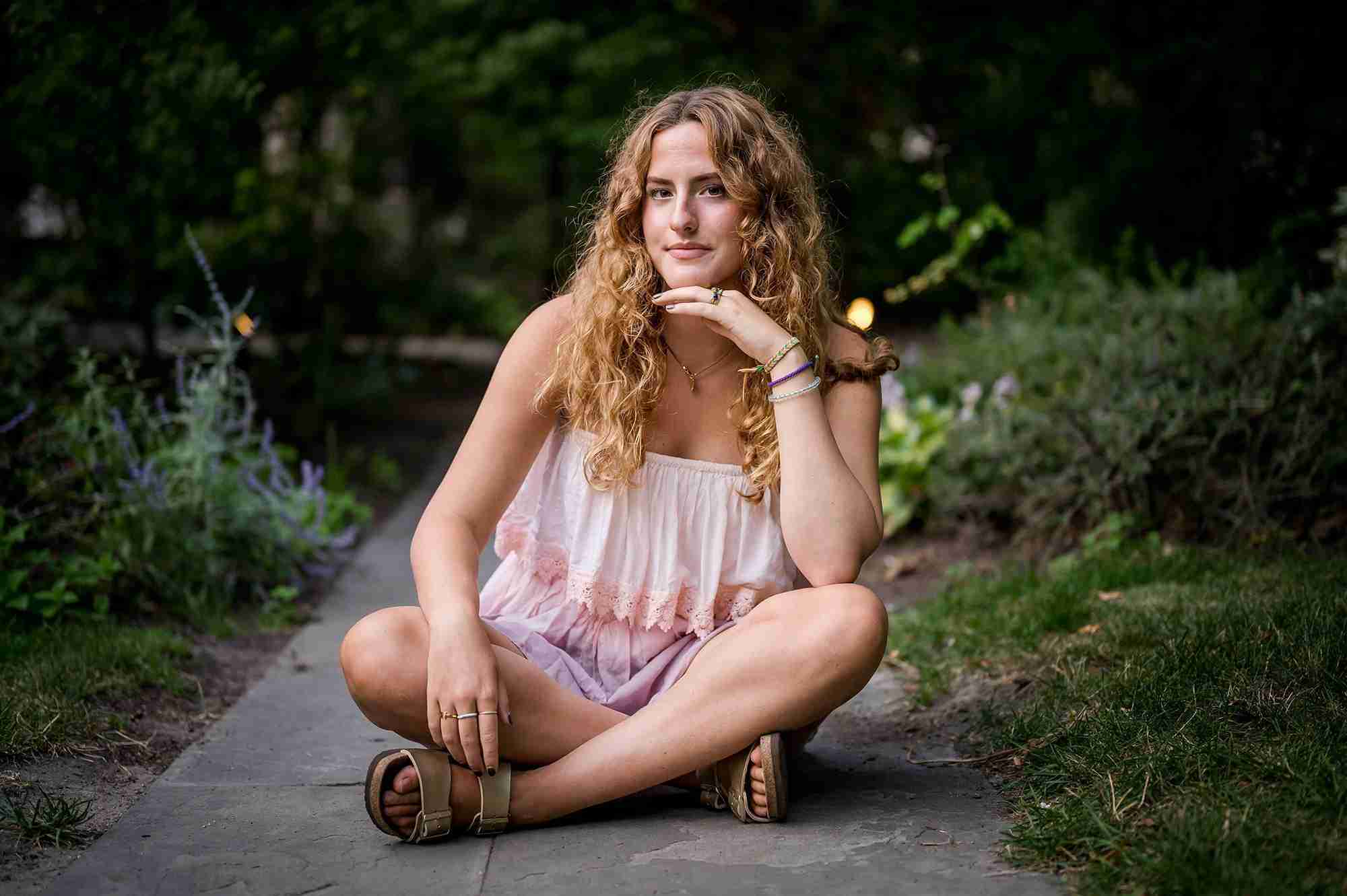 Girl in a pink strapless dress sitting with crossed legs on the sidewalk, with plants on either side of her. She's resting her elbow on her knee and her chin on her hand.