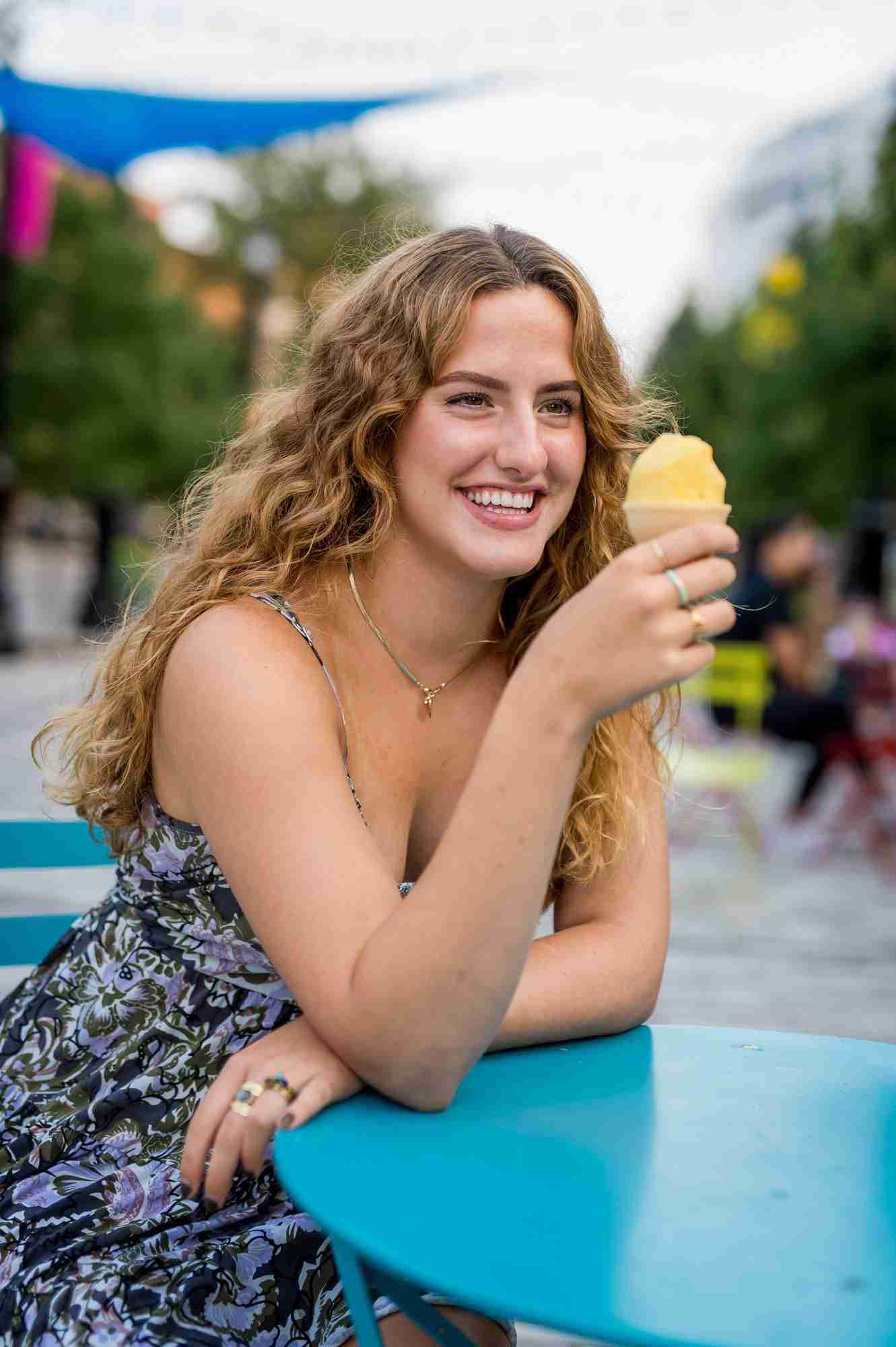 Girl wearing a blue patterned dress leaning on a blue table, holding an ice cream cone with her right hand, smiling.