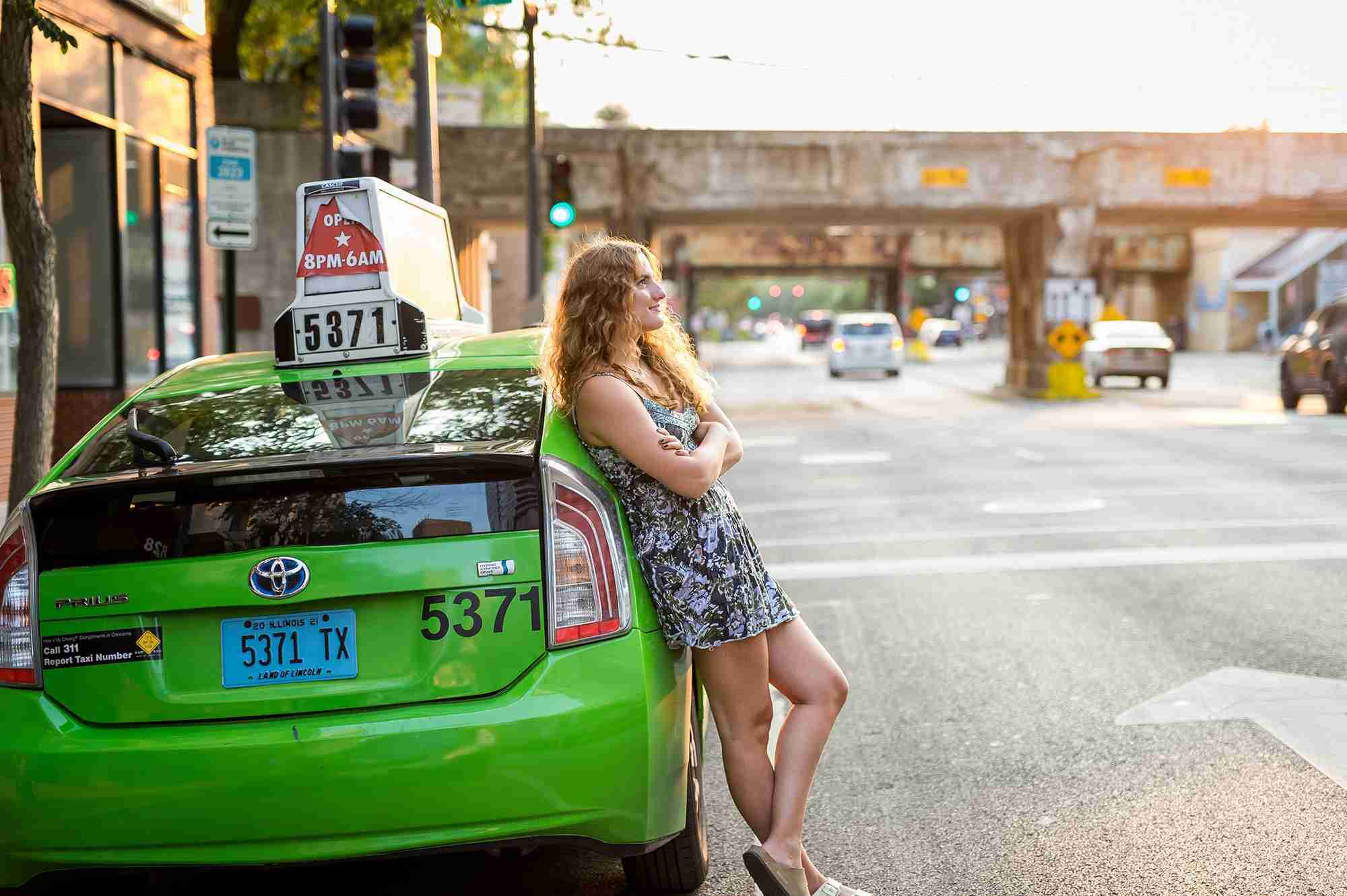Girl wearing a blue patterned dress leaning on a green taxi cab, with her legs crossed, looking straight ahead. The sun is lighting up her hair.