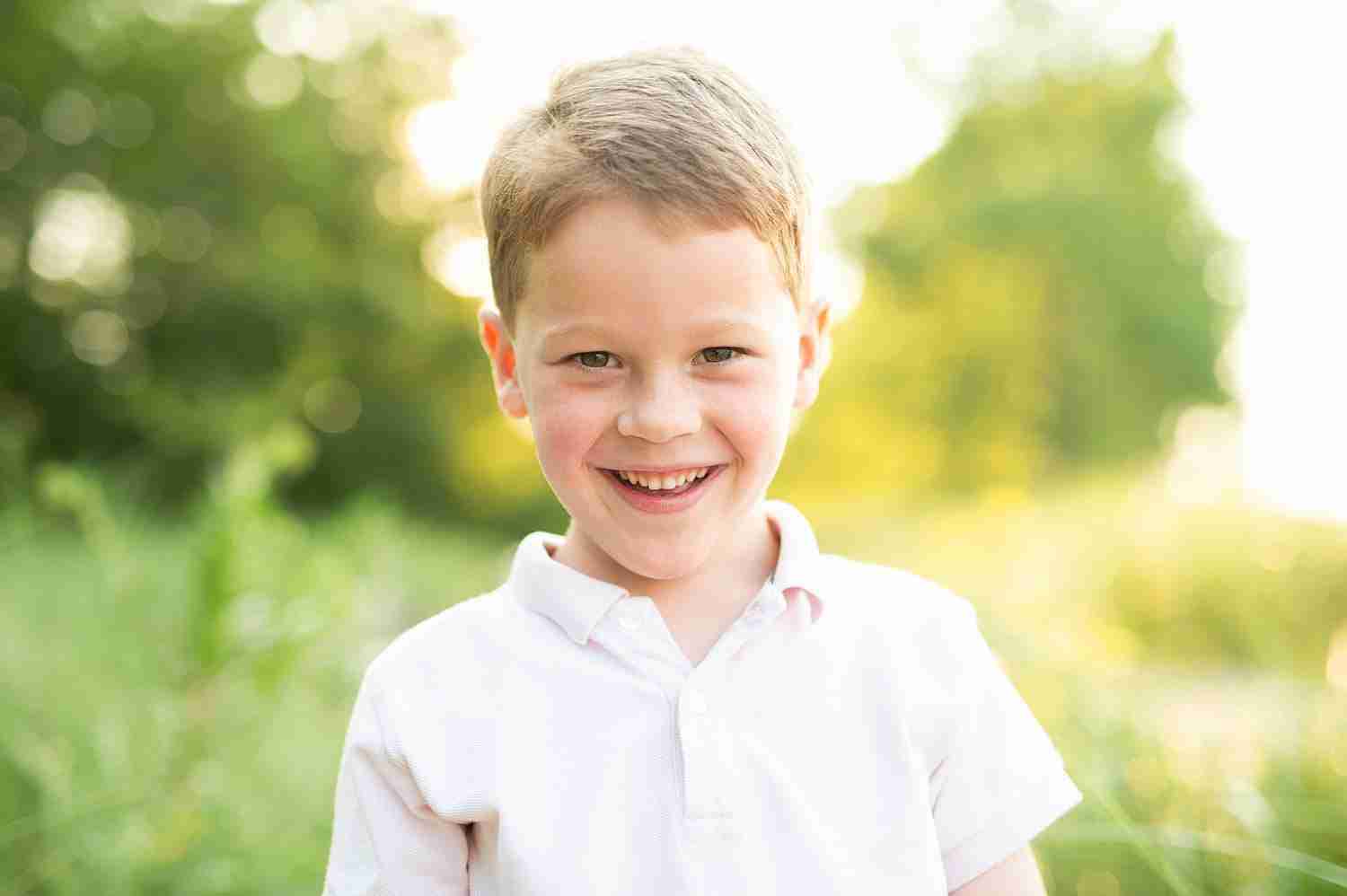 Little boy smiling with grass and trees behind him lit up by the sun.