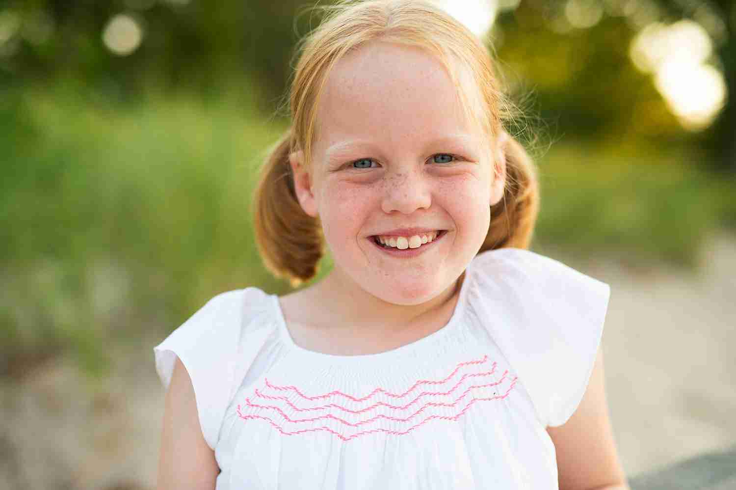Little girl with red hair and freckles smiling. She's wearing a white dress and her hair is in pigtails.