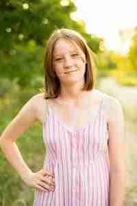 A teenage girl with shoulder length straight hair with a slight smile on her face. She's standing with her right hand on her hip and her left hand by her side. She's wearing a dress with pink and white stripes.