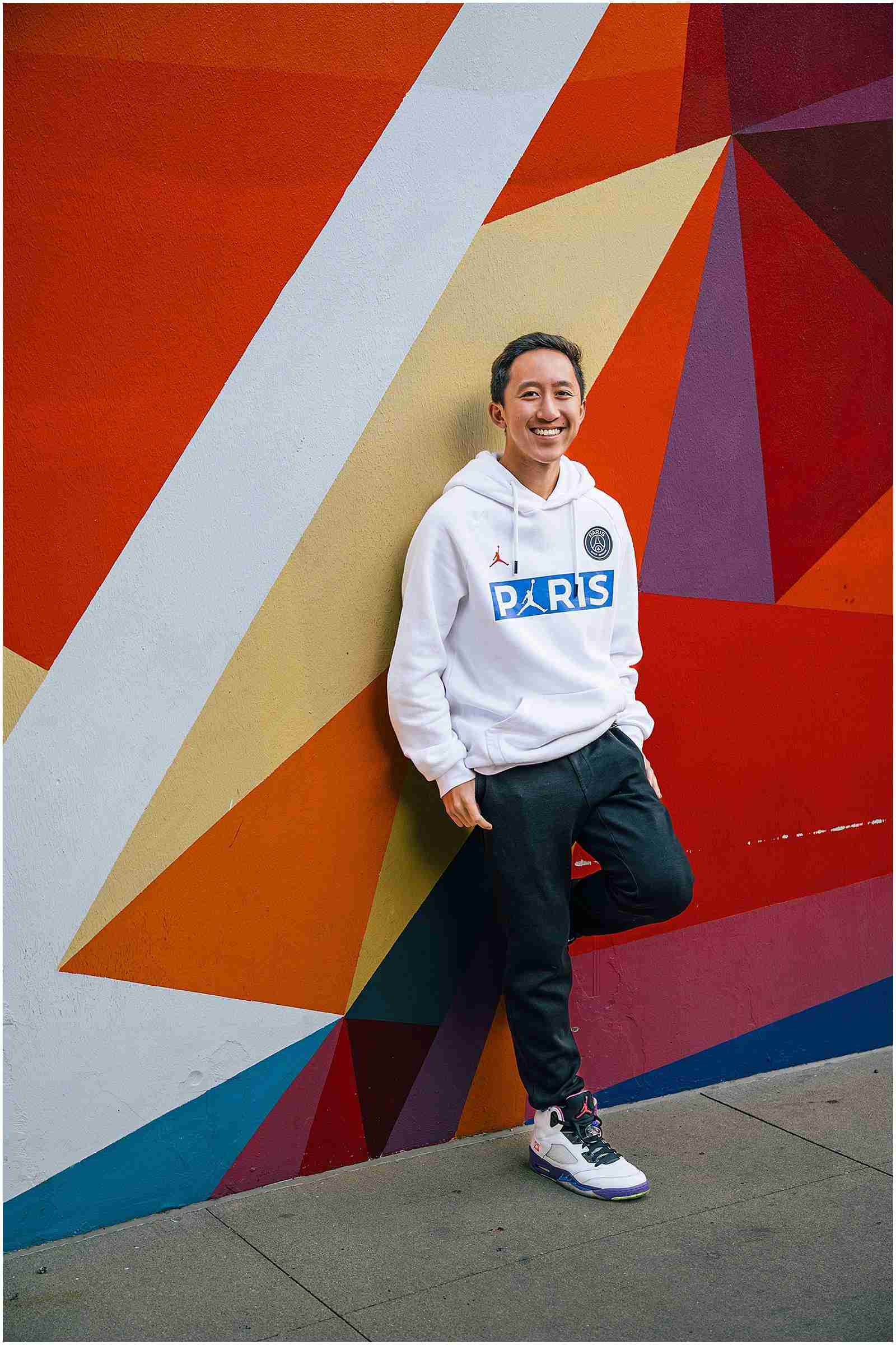 What you can expect at every senior session with SBP - senior boy smiling wearing white sweatshirt, dark jeans, and sneakers leaning against colorful mural in Chicago - award-winning Chicago senior photographer Shalimar B. Photography.