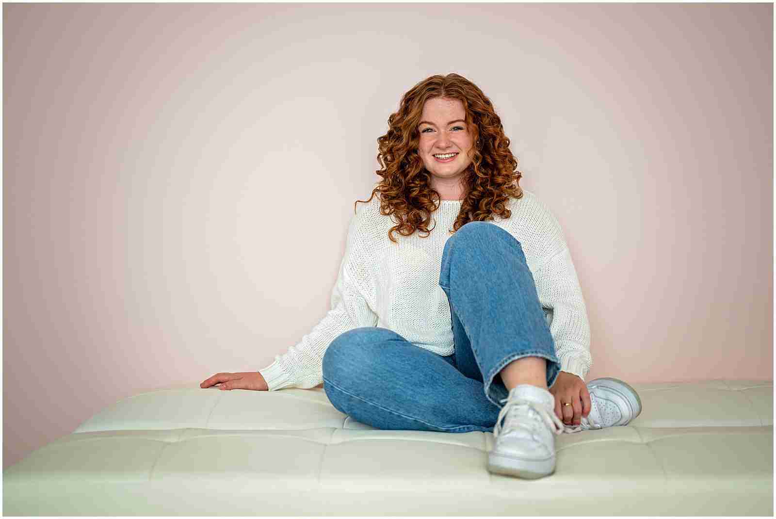 How to Book Your Senior Photos with Shalimar B. Photography with award-winning Chicago senior photographer Shalimar B. Photography. Chicago senior photos in studio of high school senior girl with red curly hair sitting on white bench wearing jeans, sneakers, and a white shirt. 
